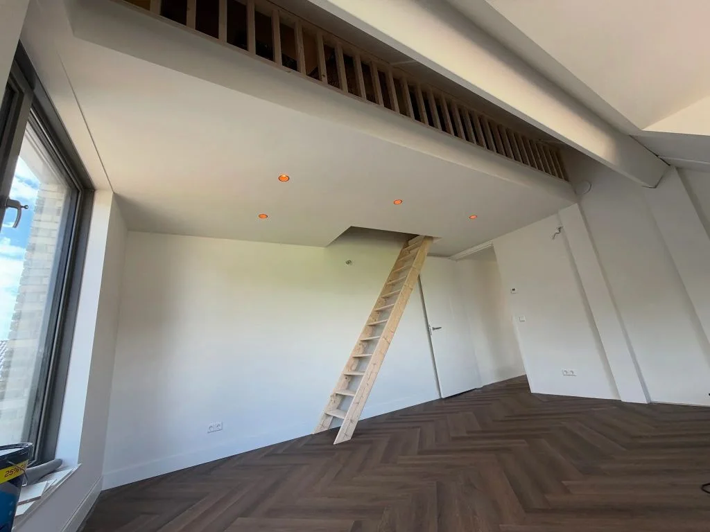 Vliering met houten balustrade en vaste trap boven een kamer met visgraatvloer